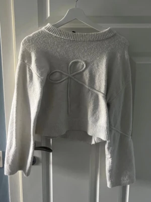 Vit stickad tröja med rosett - Superfin vit stickad tröja med en dekorativ rosett framtill. Tröjan har breda ärmar och ribbade muddar vid hals, ärmslut och nederkant. Perfekt för en trendig och mysig look.💕