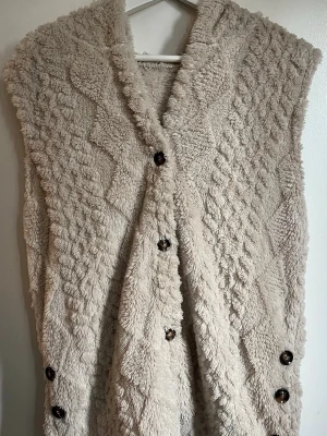 Beige fluffig pälsväst med huva - Mysig beige pälsväst med huva och dekorativa knappar framtill. Västen har ett fluffigt, mjukt material och ett stickat mönster som ger en cool struktur. Perfekt för lager på lager och för dig som gillar en avslappnad men trendig stil.