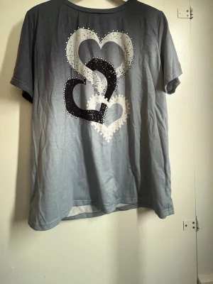 Grå t-shirt med hjärtmotiv - Snygg grå t-shirt med tre stora hjärtan i svart, vitt och grått på framsidan. T-shirten har rund halsringning och korta ärmar. Materialet är mjukt. Perfekt för en avslappnad och trendig look.