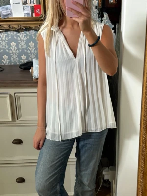 Vit plisserad ärmlös blus - Snygg vit blus med plisserat tyg och v-ringning. Blusen är ärmlös och har en lös, luftig passform som ger en avslappnad men stilren look. Perfekt att matcha med jeans eller kjol för en trendig outfit.