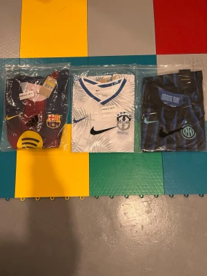 Tre fotbollströjor - Säljer tre fotbollströjor: en vinröd och blå FC Barcelona-tröja med klubbmärke och sponsorlogga, en vit och blå Brasilien-tröja från Nike med mönster och klubbmärke, samt en svart och blå Inter-tröja från Nike med klubbmärke och ränder. Alla är kortärmade och i sportigt material. Inter och Barcelona tröjan är i S och Brasilien tröjan i M. 250kr styck 500kr för alla! Kontakta vid köp