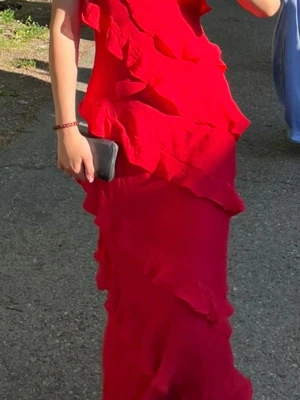 Röd långklänning med volanger - Säljer en röd långklänning med volangdetaljer och smala axelband. Klänningen har en v-ringad urringning och en figurnära passform som ger en elegant siluett. Perfekt för speciella tillfällen när du vill sticka ut. Använd endast en gång. Kan tänka mig sänka priset 