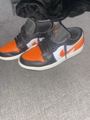 Air Jordan 1 Low Orange/Black/White - Säljer ett par Air Jordan 1 Low med orange, svart och vit färgkombination. Skorna har klassisk låg siluett, svart läder runt tån och snörning, orange tåbox och swoosh samt vit sidopanel. Ikoniska Jordan-loggor på plös och häl. Perfekta för dig som gillar streetwear.
