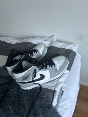 Nike Air Jordan 1 Mid grå/vit/svart - Säljer ett par Nike Air Jordan 1 Mid sneakers i grått, vitt och svart. Klassisk high-top modell med svart swoosh och svarta skosnören. Ovandel i läder och syntet, ikonisk Air Jordan-logga på sidan. Perfekta för dig som gillar streetwear och snygga detaljer.