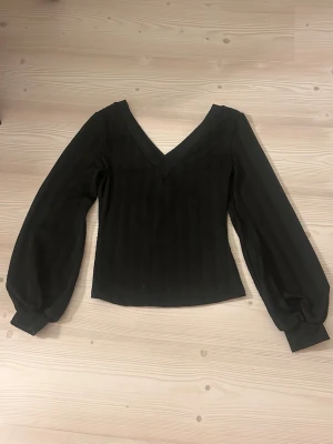 Svart v-ringad topp med ballongärmar - Snygg svart topp med v-ringning och långa ballongärmar. Toppen har en croppad passform och är tillverkad i ett lätt, något transparent material som ger en trendig look. Perfekt att styla med höga jeans eller kjol.