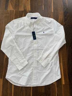 Ralph Lauren Skjorta - Klassisk vit skjorta från Ralph Lauren med blå broderad logga på bröstet. Skjortan har lång ärm, knappar framtill och button-down krage. Tillverkad i bomull för en skön och stilren känsla.