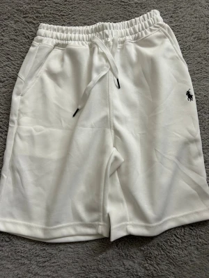 Vita shorts från Polo Ralph Lauren - Snygga vita shorts från Polo Ralph Lauren med klassisk broderad logga i svart på ena benet. De har elastisk midja med snörning och är tillverkade i mjuk bomull, perfekta för en avslappnad stil under varmare dagar.
