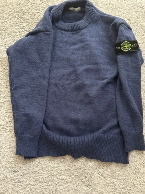 Blå stickad tröja från Stone Island - Snygg blå stickad tröja från Stone Island med klassisk rund hals och ribbade muddar. Tröjan har den ikoniska Stone Island-loggan på vänster ärm. Perfekt för dig som gillar streetwear och vill ha något stilrent och bekvämt.