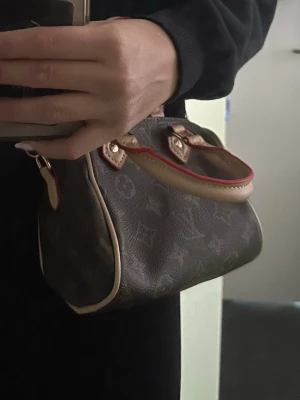 Monogram Mini Speedy - Säljer Mini Speedy handväska i klassiskt brunt monogrammönster. Väskan har ljusa läderhandtag med röda detaljer och guldfärgade metalldelar. Tillverkad i canvas med läderdetaljer och avtagbar axelrem. Perfekt för dig som gillar lyxiga accessoarer.