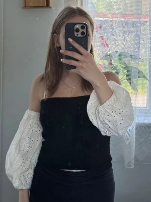 Svart topp med broderade puffärmar - Trendig svart topp med croppad passform och vita broderade puffärmar i bomull. Toppen har offshoulder-design och är perfekt för dig som vill sticka ut med en cool kontrast mellan svart och vitt. Snygg detalj med broderade mönster på ärmarna.