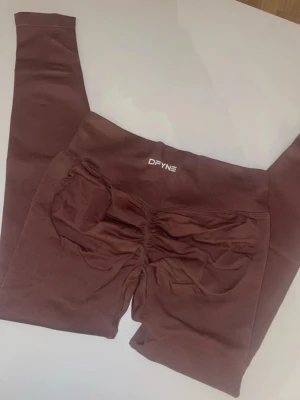 Bruna scrunch leggings från DFYNE - Snygga bruna leggings från DFYNE med scrunch-detalj bak som framhäver formerna. Hög midja och stretchigt material som sitter skönt på kroppen. Perfekta för gymmet eller chill dagar. Logga framtill i vitt.