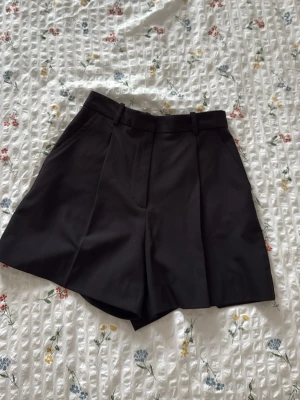 Svarta shorts från ZARA - Stilrena svarta kostymshorts från ZARA med hög midja, pressveck och hällor för skärp. Shortsen har fickor i sidorna och är tillverkade i ett slätt tyg som ger en dressad look. Perfekta för en trendig och snygg outfit.