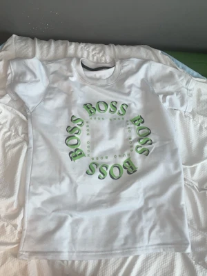 Hugo Boss set med t-shirt och shorts - Snyggt set från Hugo Boss med en vit t-shirt med grönt BOSS-tryck i cirkel på bröstet och matchande svarta shorts med liten grön logga. Perfekt för en avslappnad och trendig look. Materialet känns mjukt och bekvämt mot huden.