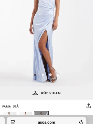 Blå långklänning med slits från ASOS - Elegant ljusblå långklänning från ASOS med omlott-design och hög slits framtill. Klänningen har öppen rygg med knytband och är tillverkad i ett glansigt material som ger en lyxig känsla. Perfekt för dig som vill sticka ut med en snygg och modern look.