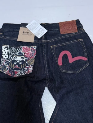 Evisu Selvedge Limited - Säljer ett par extremt sällsynta och helt oanvända jeans från japanska Evisu. Detta är Lot 2010-modellen, vilket innebär en ”Slim Straight”-passform. Den är något smalare över låren och benen jämfört med den klassiska 2008-modellen, vilket ger en modern och skarp siluett. NYPRIS—>3600kr HELT OANVÄNDA🤩 Mått:   • Midja (rakt över): 38 cm • Höft: 53 cm • Ytterben (total längd): 107 cm • Innerben (gren till benslut): 80 cm • Lår: 22 cm • Benslut (bredd): 18 cm MÅTT KAN VARA +/-1cm 