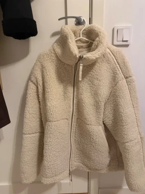 Beige teddyjacka med dragkedja - Mysig beige teddyjacka med hög krage och hel dragkedja framtill. Jackan har två stora fickor och är tillverkad i fluffigt teddy-material som håller dig varm. Perfekt för kyliga dagar och enkel att matcha med olika outfits. Använd knappast minst 2-3 gånger
