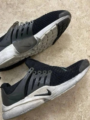 Nike Air Presto svarta sneakers - Nike Air Presto sneakers i svart mesh med grå detaljer och vit sula. Skorna har en sportig design med plastdetaljer på sidorna och klassisk Nike-logga. Ovandelen är luftig och flexibel, perfekt för en aktiv livsstil.
