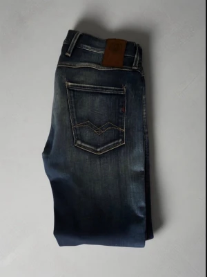 Mörkblå slim fit jeans från Replay - Snygga mörkblå slim fit jeans från Replay med slitningar framtill och klassiska fem fickor. Jeansen har kontrastsömmar och en läderpatch bak i midjan. Tillverkade i bomull med en liten mängd stretch för extra komfort. Perfekt till sommarn