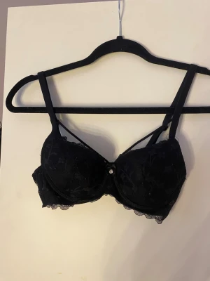 Svart spets-bh med strassdetalj - Snygg svart bh med spetsdetaljer. Dekorativ strassdetalj mellan kuporna och extra band över bysten för en trendig look. Bh:n har justerbara axelband och fina spetskanter. Endast testad inte använd