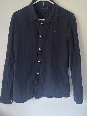 Mörkblå skjorta från Tommy Hilfiger - Klassisk mörkblå skjorta från Tommy Hilfiger med diskret logga på bröstet. Skjortan har långa ärmar, knappar framtill och en stilren krage. Perfekt för en clean och enkel look.