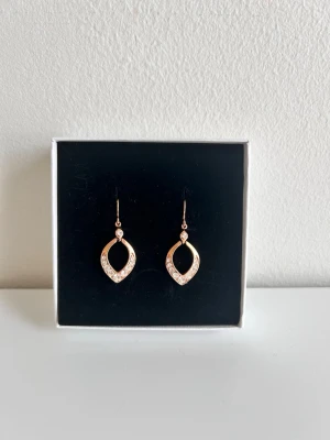 Guldiga örhängen med gnistrande stenar - Ett par Pilgrim örhängen i guldton med droppformad design. Örhängena har små gnistrande stenar längs kanten som ger extra bling. Perfekta för dig som vill ha något stilrent men ändå iögonfallande. Krokar i matchande färg.