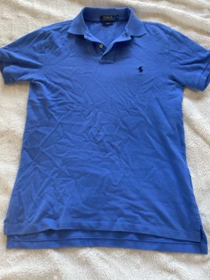 Blå pikétröja från Ralph Lauren - Klassisk blå pikétröja från Ralph Lauren med den ikoniska broderade loggan på bröstet. Tröjan har krage, knappar vid halsen och korta ärmar. Perfekt för en avslappnad men ändå stilren look, och även perfekt för sommaren 