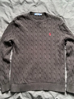 Grå kabelstickad tröja från Polo Ralph Lauren - Kabelstickad grå tröja från Polo Ralph Lauren med rund halsringning och ribbade muddar. Tröjan har en klassisk passform och är dekorerad med det ikoniska röda Polo-logot på bröstet. Perfekt för lager-på-lager och stilrena outfits.