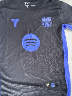 FC Barcelona svart matchtröja Nike - Snygg svart FC Barcelona matchtröja från Nike med lila detaljer och klubbmärke på bröstet. Tröjan har korta ärmar, lila sidopaneler och tryck av Spotify-loggan på framsidan. Tillverkad i lätt och ventilerande funktionsmaterial, perfekt för fotbollsträning.