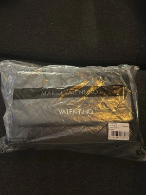 Svart Valentino Marion väska - Snygg svart axelväska från Mario Valentino med quiltat mönster och silverfärgad kedjerem. Väskan har en stilren rektangulär form och logga framtill. Används max 2 gånger snabbt då de va spontanköp och har andra väskor. Nypris 2000kr WAKANDA modellen. Du får med orginalpåse