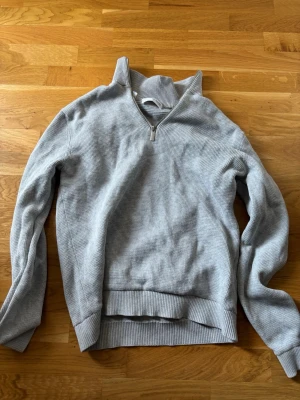 Grå halvzip tröja Selected Homme - Snygg grå tröja från Selected Homme med halv dragkedja och hög krage. Tillverkad i mjukt stickat material med ribbade muddar vid ärmslut och nederkant. Perfekt för lager på lager och enkel att matcha med jeans eller chinos.