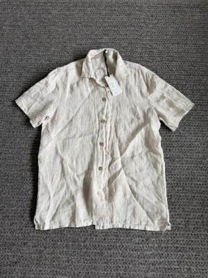 Beige linneskjorta från Befree - Snygg beige skjorta från Befree i linne med korta ärmar och klassisk krage. Skjortan har knappar framtill och en avslappnad passform, perfekt för varma dagar.