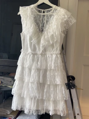 Vit spetsklänning med volanger - Superfin vit kort klänning i spets med flera lager volanger och v-ringning både fram och bak. Klänningen har ärmlös design och knäppning i nacken. Perfekt för dig som gillar romantisk och feminin stil. Ifrån bubbleroom perfekt till studenten 