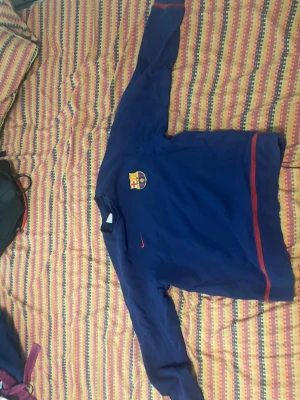 Blå FC Barcelona tröja Nike - Mörkblå långärmad tröja från Nike med FC Barcelona-logga på bröstet och röda detaljer vid ärmslut och nederkant. Perfekt för fotbollsfans som vill visa sitt stöd för laget. Tröjan har en sportig och bekväm passform.