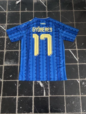 Sverige bortaställ VM 2026 | Gyökeres #17 - Säljer en blå Sverige fotbollströja från Adidas med gult tryck, nummer 17 och namnet Gyökeres på ryggen. Tröjan har korta ärmar, gula ränder på axlarna och Sveriges landslagsemblem på bröstet. Snyggt mönster i blått över hela tröjan.