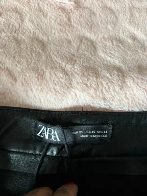 Svart skinnkjol från Zara XS - Snygg svart kort skinnkjol från Zara i storlek XS. Kjolen har bälteshällor och en stilren design som ger en cool vibe. Perfekt att matcha med en stickad tröja eller t-shirt för en trendig look.