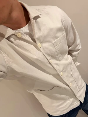 Vit overshirt  - Stilren vit overshirt från Weekday med klassisk krage, stora knappar och tre fickor framtill. Skjortan är långärmad och tillverkad i bomull, perfekt att bära öppen över en t-shirt för en avslappnad look.