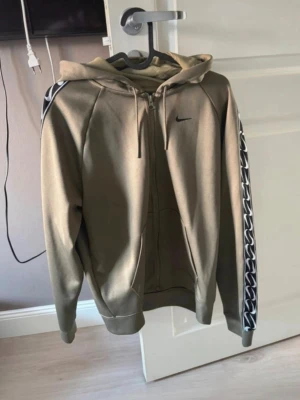 Nike hoodie med dragkedja och mönstrade ärmar - Snygg olivgrön hoodie från Nike med dragkedja och huva. Ärmarna har ett svartvitt mönster längs sidorna och en liten svart Nike-logga på bröstet. Perfekt för en sportig och avslappnad stil.