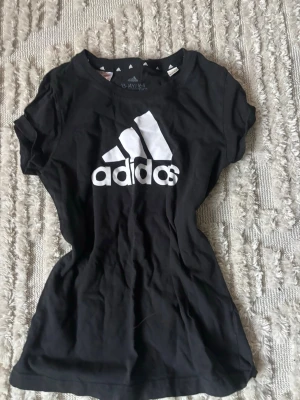 Svart Adidas t-shirt med logga - Svart t-shirt från Adidas med stor vit logga på bröstet. Klassisk rund halsringning och korta ärmar. Tillverkad i mjuk bomull som känns skön mot huden. Perfekt för en sportig och avslappnad stil.