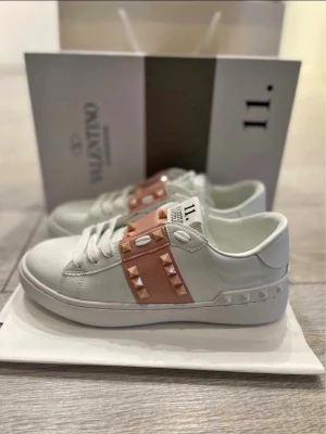 Valentino Rockstud Untitled sneakers - Snygga vita sneakers från Valentino med ljusrosa band och ikoniska nitar i samma färg. Skorna har en klassisk låg modell med rund tå och vita skosnören. Tillverkade i läder med detaljer som ger en lyxig och trendig vibe.