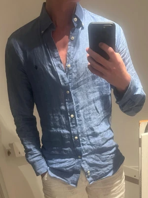 Blå linneskjorta från Morris - Snygg blå skjorta i 100% linne från Morris med klassisk button-down krage och vita knappar. Skjortan har lång ärm och en liten broderad logga på bröstet. Perfekt för en avslappnad men stilren look.