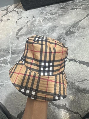 Beige bucket hat med rutigt mönster - Snygg bucket hat i beige med klassiskt rutigt mönster i svart, vitt och rött. Hatten har en mjuk, nedvikt brätte och är perfekt för en trendig streetstyle-look. Materialet känns lätt och bekvämt.