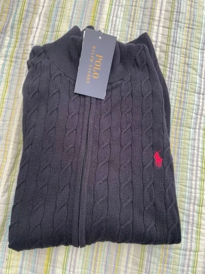 Marinblå kabelstickad tröja Polo Ralph Lauren - Svart kabelstickad tröja från Polo Ralph Lauren med dragkedja och hög krage. Tröjan har det klassiska röda broderade logotypen på bröstet och är tillverkad i mjukt material. Perfekt för en stilren och avslappnad look.