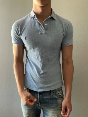 Ljusblå pikétröja från Ralph Lauren - Snygg ljusblå pikétröja från Ralph Lauren med klassisk krage och två knappar framtill. Tröjan har korta ärmar och den ikoniska loggan broderad på bröstet. Perfekt för en stilren och avslappnad look.