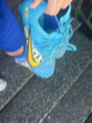Blå Nike Mercurial fotbollsskor - Säljer ett par blå Nike Mercurial fotbollsskor med gula och svarta detaljer samt tydlig Swoosh-logga på sidan. Skorna har snörning och mönstrad ovandel i syntetmaterial för bra grepp och känsla på planen. Perfekta för dig som vill sticka ut på fotbollsplanen!