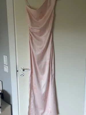 Ljusrosa långklänning med blommor - Elegant ljusrosa långklänning med tunna axelband och subtilt blommönster. Klänningen har en asymmetrisk halsringning och är figurnära med ett mjukt, glansigt material som ger en lyxig känsla. Perfekt för speciella tillfällen. Använd ca en gång