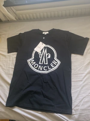 Svart Moncler t-shirt med stor logga - Svart t-shirt från Moncler med stor vit logotyp på bröstet. Klassisk rund halsringning och korta ärmar. Tillverkad i mjuk bomull som känns skön mot huden. Perfekt för dig som gillar stilrena och trendiga plagg med tydlig branding.