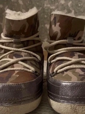 Inuikii stövlar stl 40 - Säljer ett par moon boots med camouflagemönster och metallicgrå detaljer. Skorna har grova snören och är fodrade med mjuk, vit päls på insidan. Perfekta för kalla dagar när du vill sticka ut med en cool stil.40
