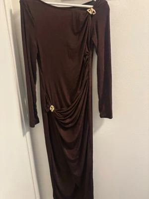 Brun långklänning med gulddetaljer - Elegant brun långklänning med långa ärmar och draperad design. Klänningen har två guldfärgade detaljer vid axeln och höften som ger en lyxig touch. Perfekt för dig som vill sticka ut med en unik och stilren look.