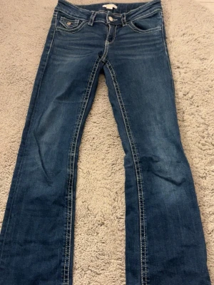 Blå bootcut jeans med kontrastsömmar - Säljer ett par klassiska blå bootcut jeans med snygga kontrastsömmar och fem fickor. Jeansen har låg midja och är tillverkade i ett mjukt denimtyg som ger en avslappnad look. Perfekta för dig som gillar en tidlös stil med lite retrovibbar.