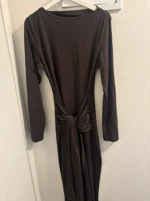 Brun långärmad klänning med knyt - Mörkbrun långklänning med långa ärmar och snygg knytning i midjan. Klänningen har en enkel, elegant design med rund halsringning och är tillverkad i ett mjukt, stretchigt material som ger en smickrande passform.